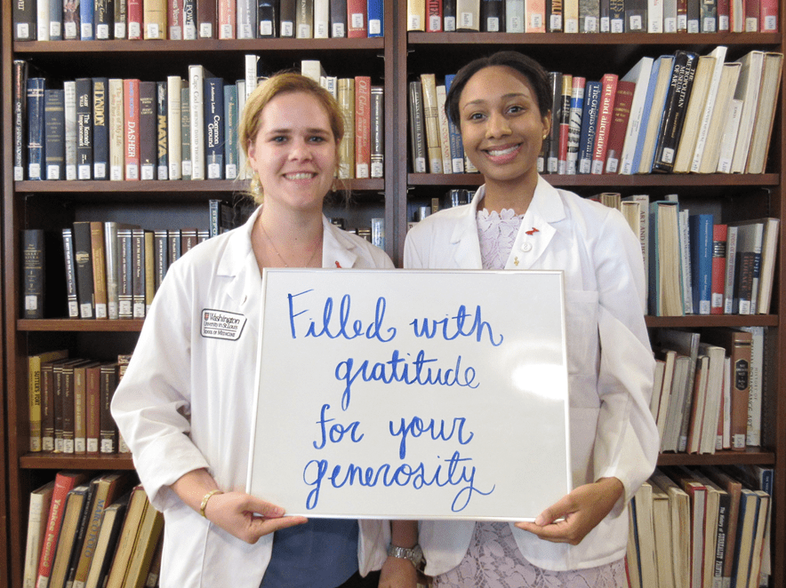 Medical students in white coats stand before a bookcase holding a sign that reads "Filled with gratitude for your generosity"
